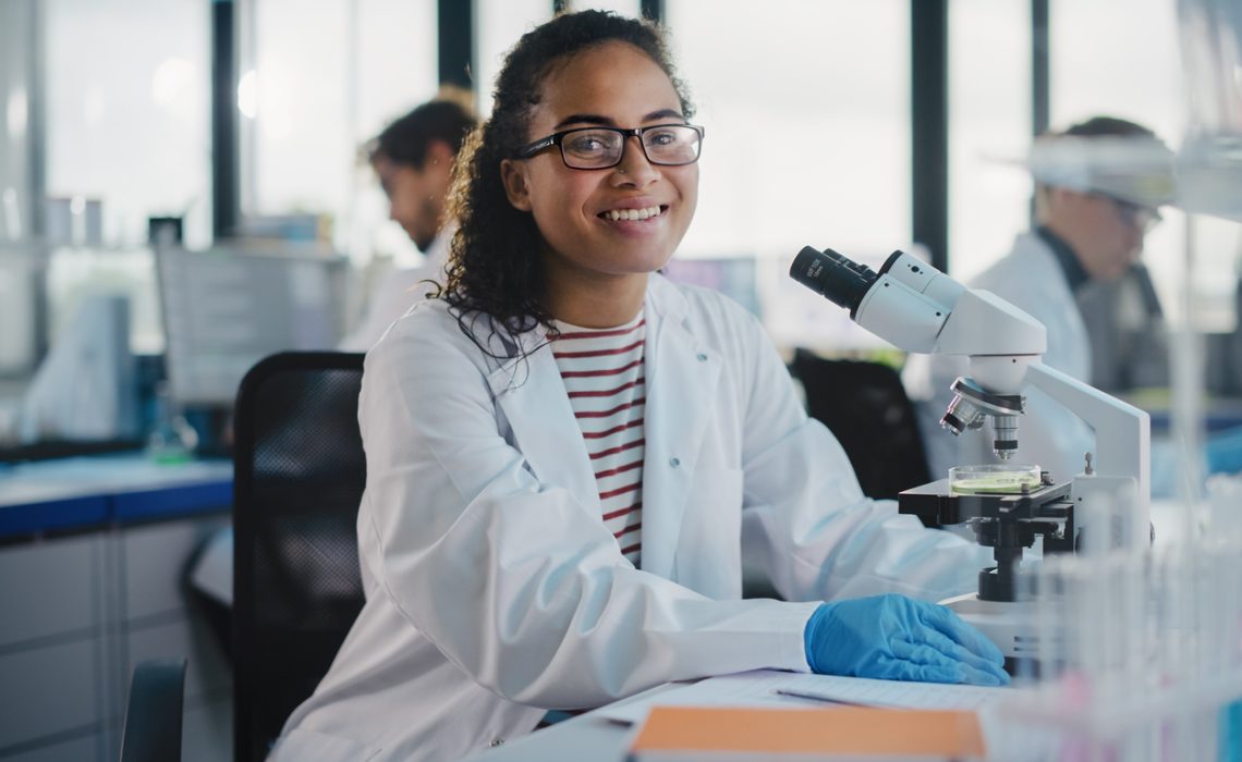 Medical Science Laboratory: Beautiful Black Scientist is Using Microscope, Looking at Camera and Smiling Charmingly. Young Biotechnology Science Specialist, Using Technologically Advanced Equipment.