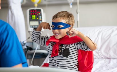 Cheerful strong little boy wearing blue eyeband and red cape like superhero sitting in hospital bed playing with nurse