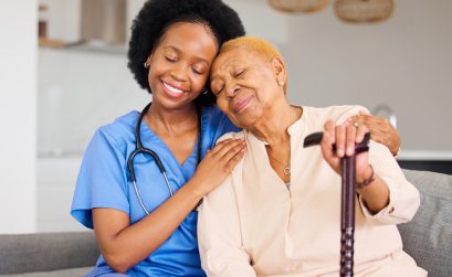 Black woman, nurse and senior patient in elderly care, hug and healthcare on living room sofa at home. Happy African medical professional or caregiver help person with kindness and cane in house