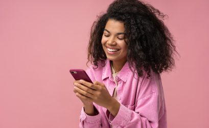 Smiling African American woman using smartphone playing mobile game isolated on pink background. Happy stylish female holding mobile phone shopping online with sale