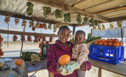 Happy African street vendor holding her child, shack with vegetables and fruits