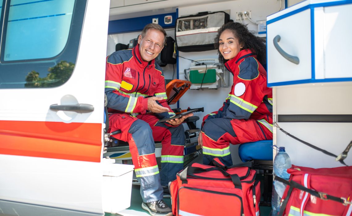 Pleased doctor with the tablet sitting beside his joyous young female colleague in the medical emergency vehicle