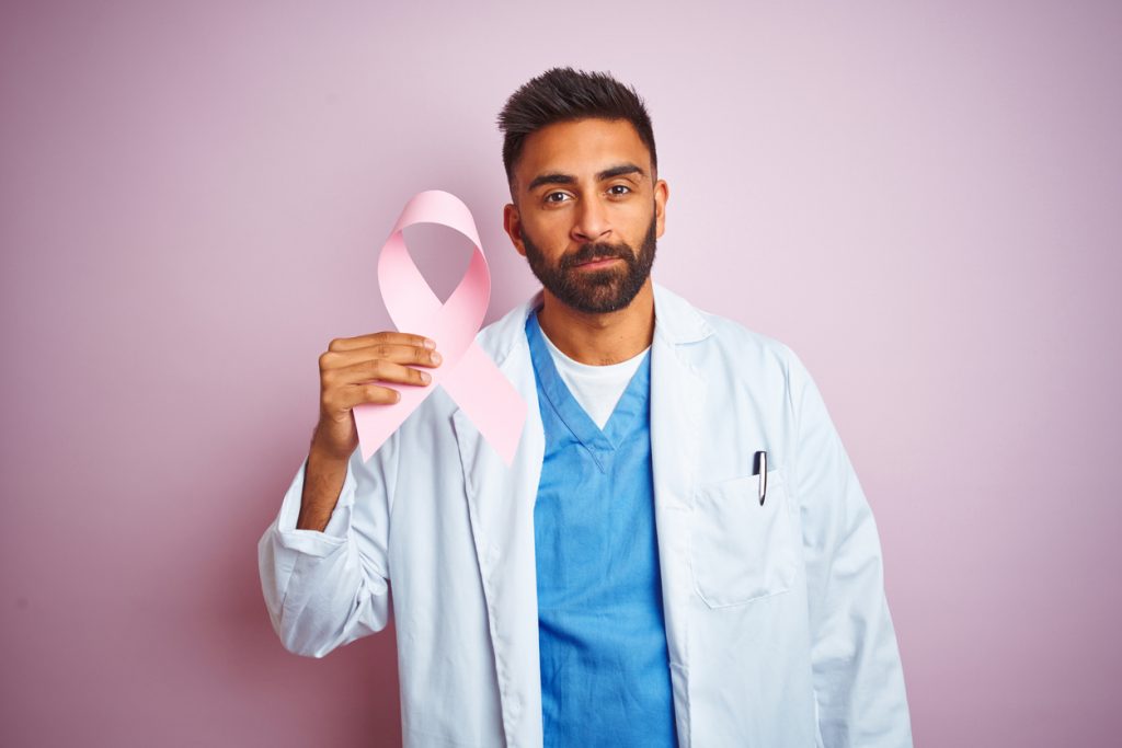 Young indian doctor man holding cancer ribbon standing over isolated pink background with a confident expression on smart face thinking serious
