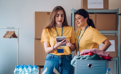 Two woman volunteers working in community charity donation center. Volunteers putting clothes and other supplies in donation boxes while social worker is making notes on a digital tablet.