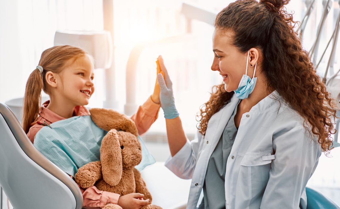 Cute little girl sitting in dentist chair, giving high five to female doctor and laughing. Dental care, trust and patient care. Children's dentistry.Sunlight.