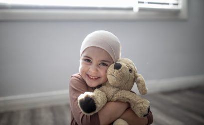 A sweet little girl battling cancer sits at home on the floor of her bedroom, with her arms wrapped tightly around her stuffed animal as she smiles gently for the portrait. She is dressed casually and wearing a headscarf as the sun shines in on her through the window.