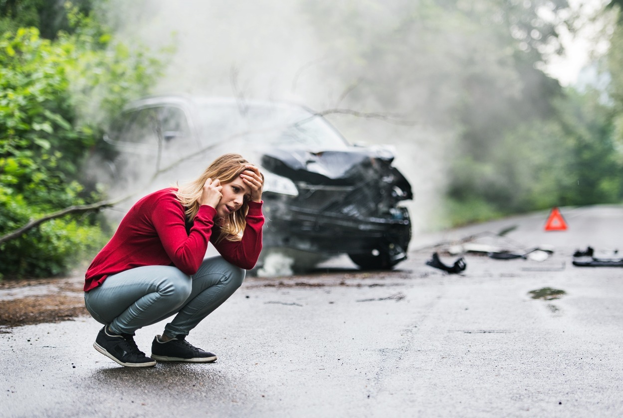 A frustrated young woman with smartphone by the damaged car after a car accident, making a phone call