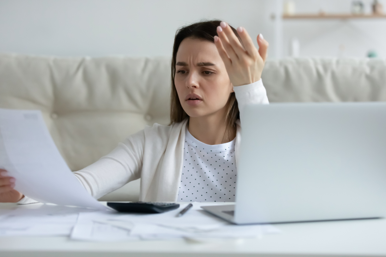 Irritated young woman checking financial documents, using laptop, online banking service, angry unhappy girl reading bad news in letter, calculating domestic bills, sitting at table at home